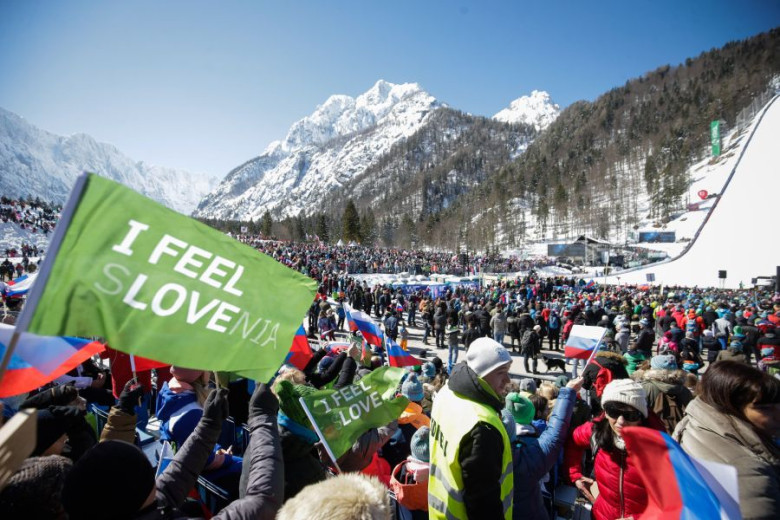 Crowd at a ski jumping event in Planica, surrounded by snowy mountains, waving Slovenian flags and banners.