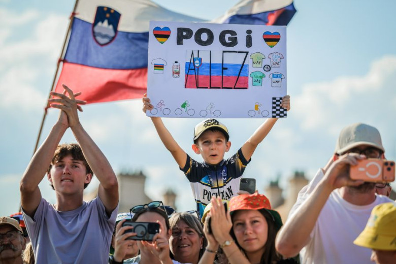 Young boy holds a sign saying "POGI Allez," surrounded by cheering crowd, Slovenian flag waving in the background, 