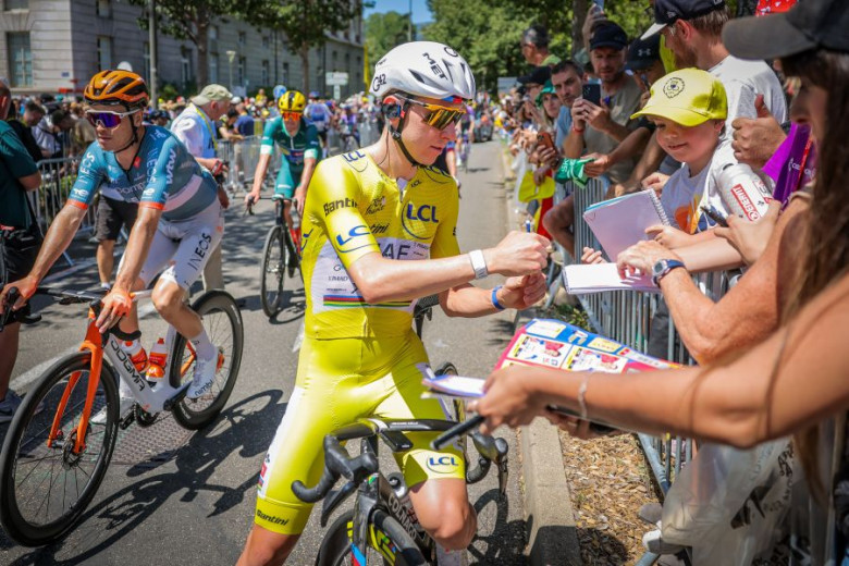 Tadej Pogačar in yellow jersey signs autographs for fans along a crowded race route. 