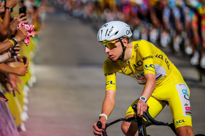 Tadej Pogačar wearing the yellow jersey on his bike, with a crowd of spectators along the roadside in the background.