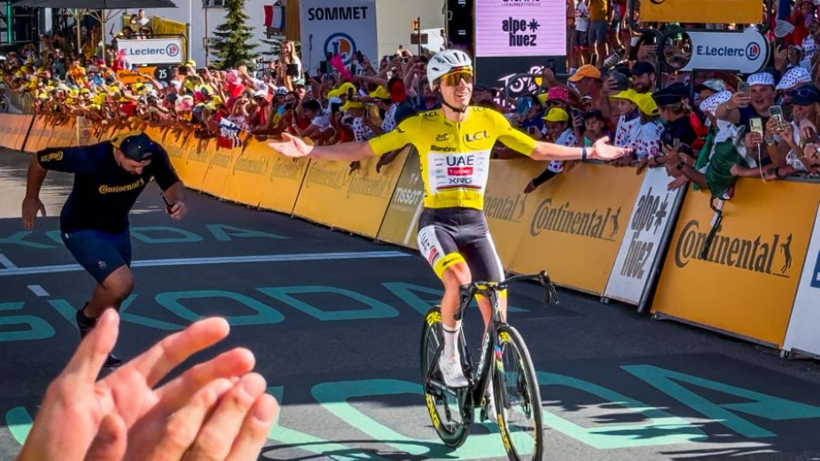Tadej Pogačar in the yellow jersey crosses the finish line with his arms outstretched in victory, as a cheering crowd lines the road.