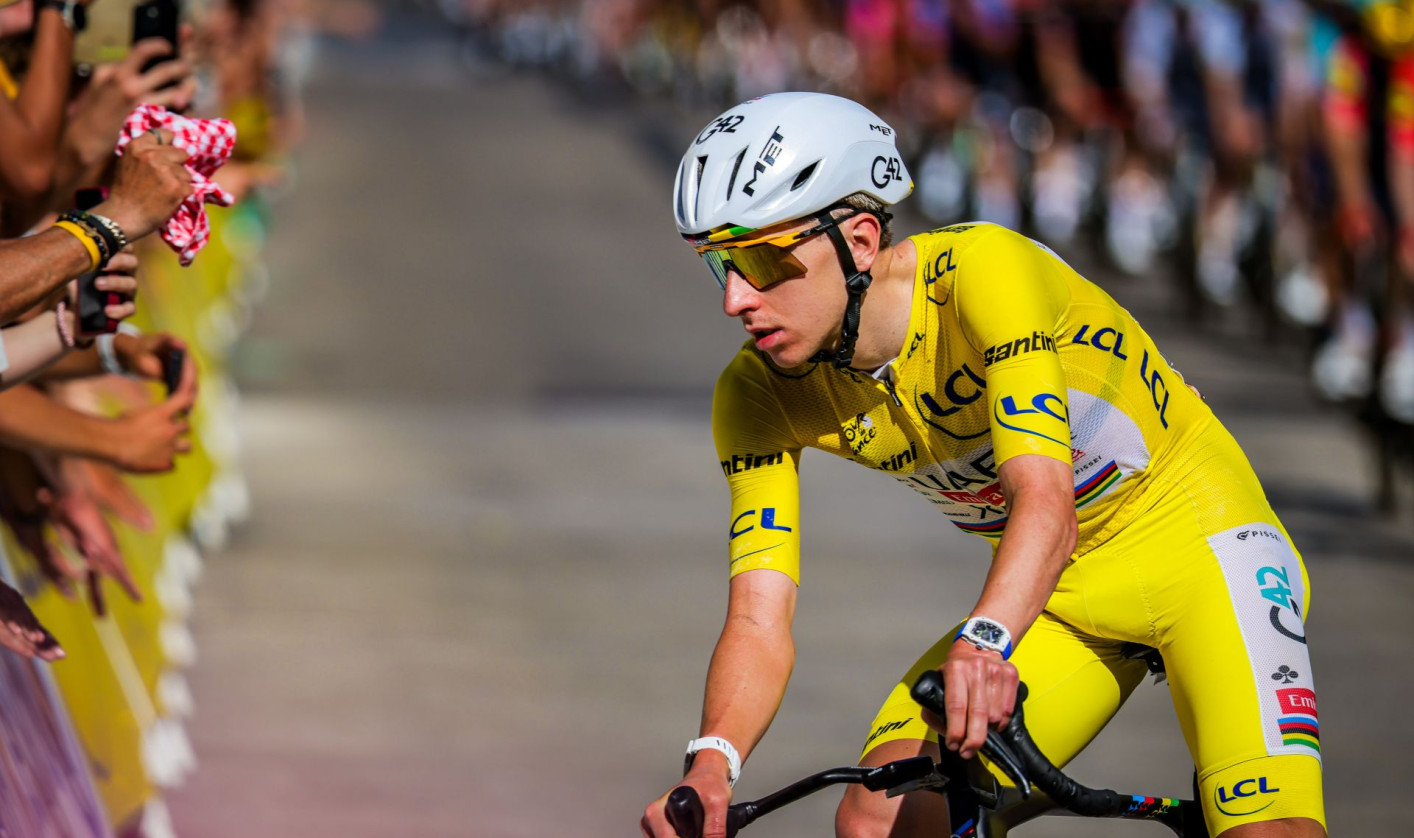 Tadej Pogačar wearing the yellow jersey on his bike, with a crowd of spectators along the roadside in the background.