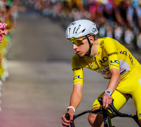 Tadej Pogačar wearing the yellow jersey on his bike, with a crowd of spectators along the roadside in the background.