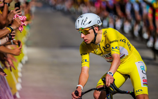 Tadej Pogačar wearing the yellow jersey on his bike, with a crowd of spectators along the roadside in the background.