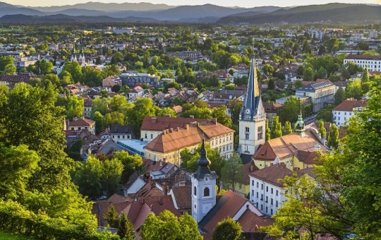 Aerial view of Ljubljana old city with a point-shaped roof church and other buildings surrounded by greenery, trees and hills in the distant background.