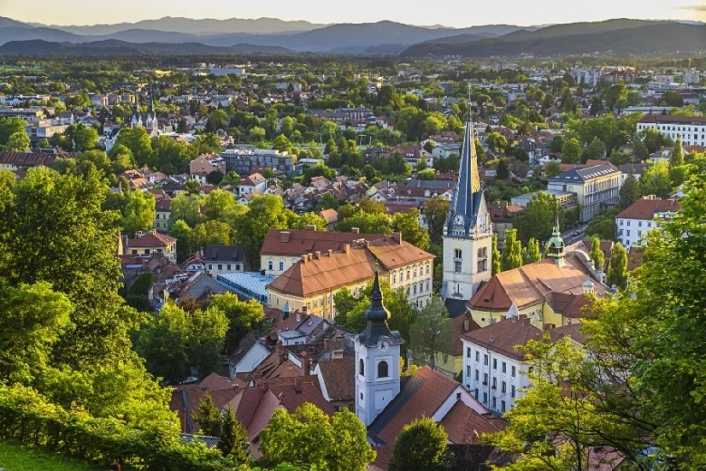 Aerial view of Ljubljana old city with a point-shaped roof church and other buildings surrounded by greenery, trees and hills in the distant background.