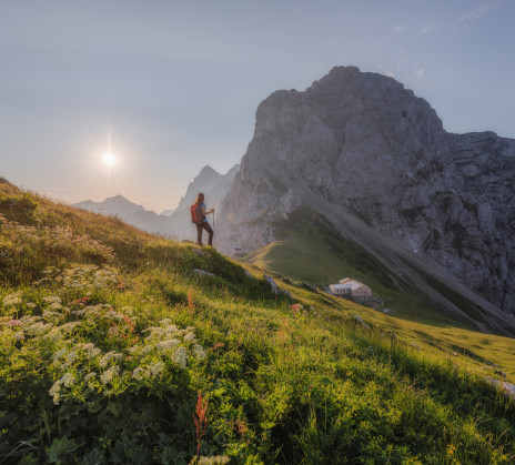 A hiker with a backpack standing on a grassy slope at sunset, overlooking steep rocky mountains and a mountain hut below.