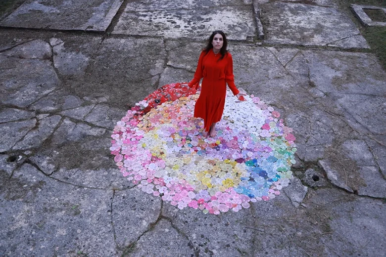 A woman in a red dress stands on a circular installation of multicolored handmade lace roses on the ground, looking upward.