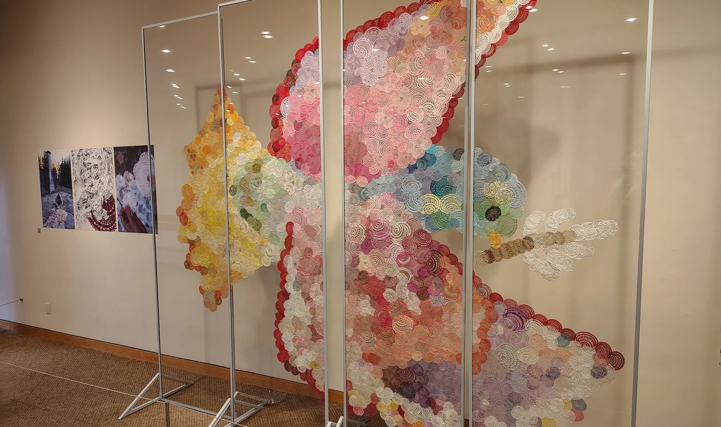 In a gallery space, several transparent panels display a large art installation in the shape of a bird. It is composed of numerous multicolored handmade lace patterns that together form the image of a bird in flight.