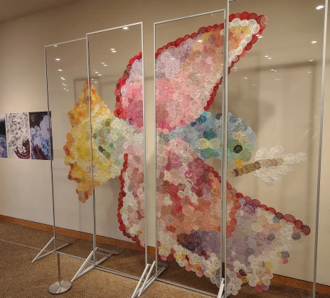 In a gallery space, several transparent panels display a large art installation in the shape of a bird. It is composed of numerous multicolored handmade lace patterns that together form the image of a bird in flight.