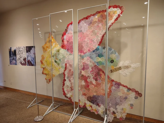 In a gallery space, several transparent panels display a large art installation in the shape of a bird. It is composed of numerous multicolored handmade lace patterns that together form the image of a bird in flight.