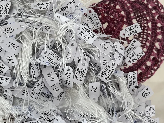 A bundle of white threads with attached tags bearing handwritten names of the creators of handmade lace roses.