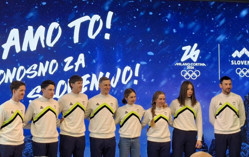 Skupina športnikov v belih reprezentančnih puloverjih stoji drug ob drugem na odru, nasmejani, za njimi modro ozadje z logotipi OI 2026.