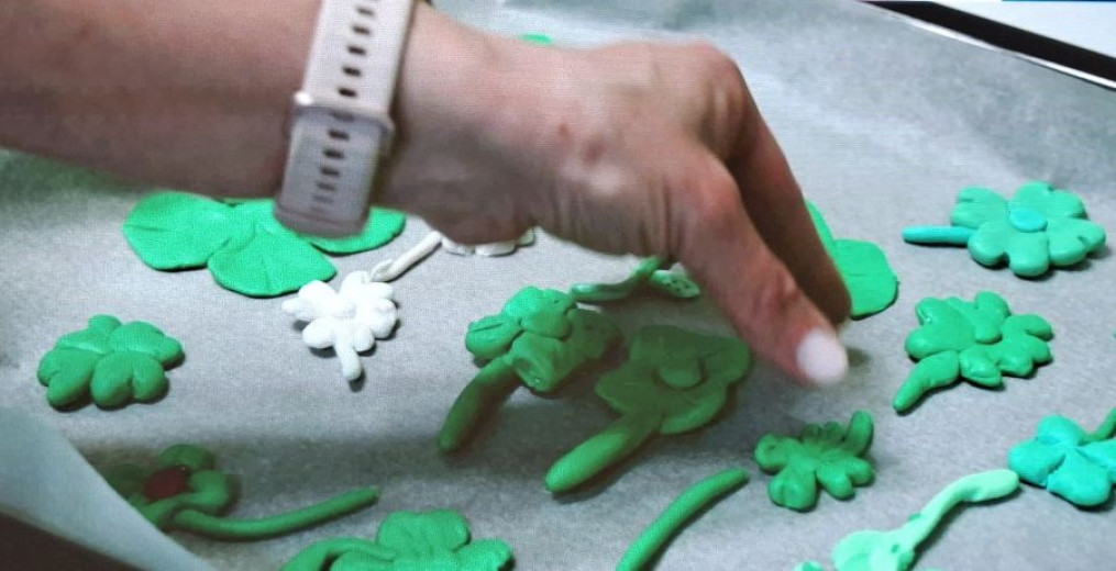 Roka z belo uro razporeja zelene testene oblike, podobne štiriperesnim deteljicam, na pekač, obložen s papirjem za peko, kar vzbuja občutek ustvarjalnosti in zbranosti.