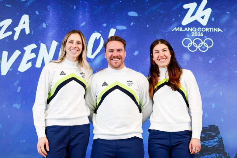 Trije ljudje v ujemajočih se belih in modrih uniformah se smehljajo pred ozadjem Milano Cortina 2026. Vzdušje je veselo in praznično.