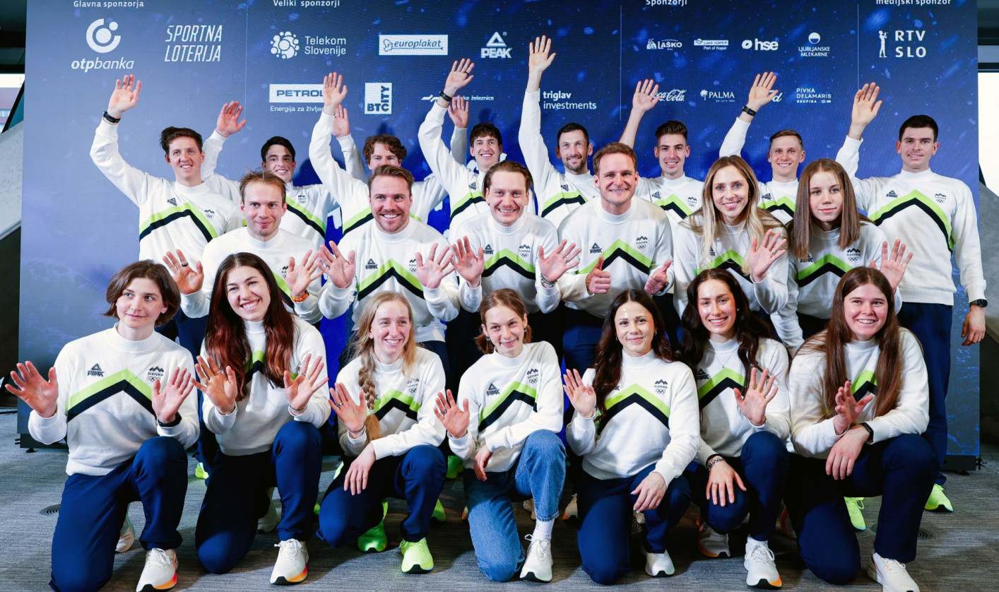 A group of people in matching white and green sportswear smiles and waves at the camera. They stand in front of a sponsor-covered backdrop, conveying a cheerful and team-oriented atmosphere.