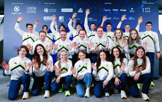 Skupina ljudi v ujemajočih se belih in zelenih športnih oblačilih se smehlja in maha proti kameri. Stojijo pred ozadjem, prekritim z logotipi sponzorjev, kar ustvarja veselo in ekipno vzdušje.