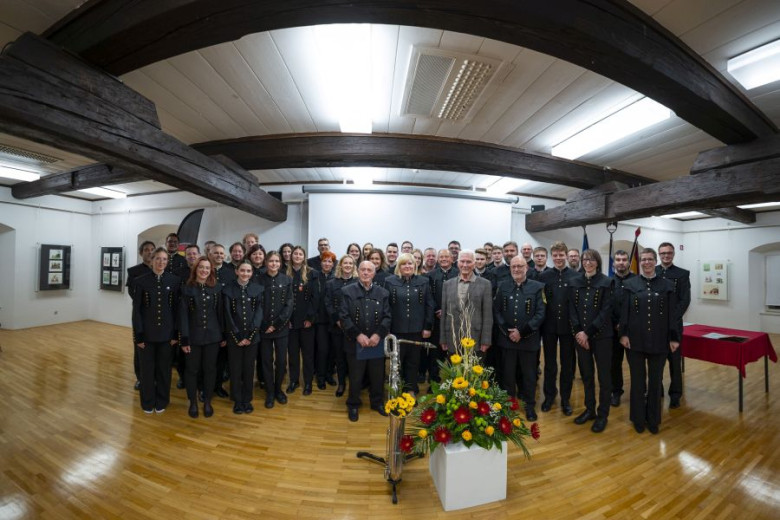 Group photo of a uniformed band indoors; wooden floor and beams; flower arrangement in front.