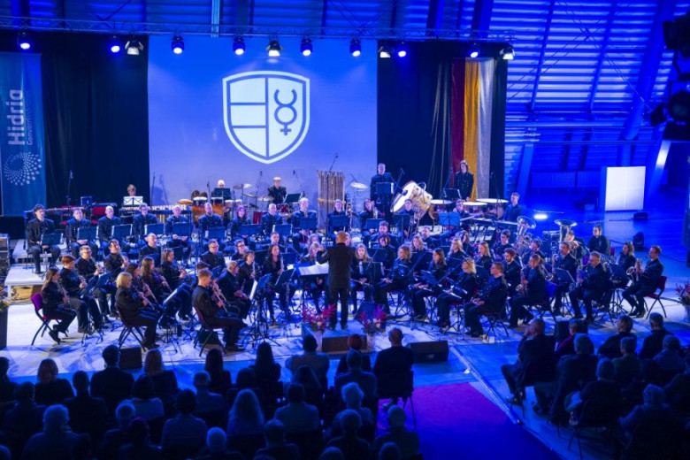 Large symphonic orchestra on stage under blue lights; conductor centered; audience seated in foreground.