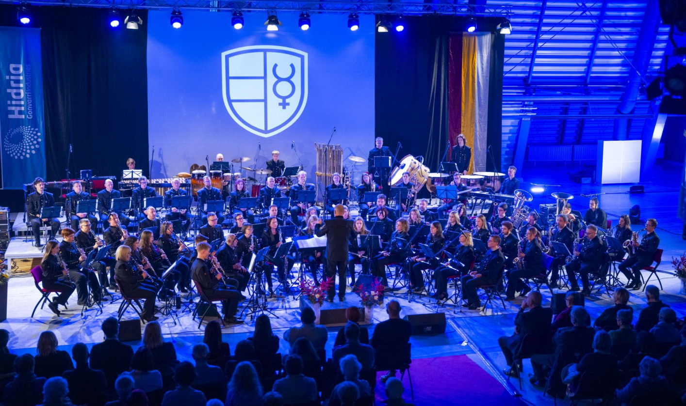 Large symphonic orchestra on stage under blue lights; conductor centered; audience seated in foreground.