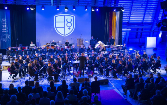 Large symphonic orchestra on stage under blue lights; conductor centered; audience seated in foreground.