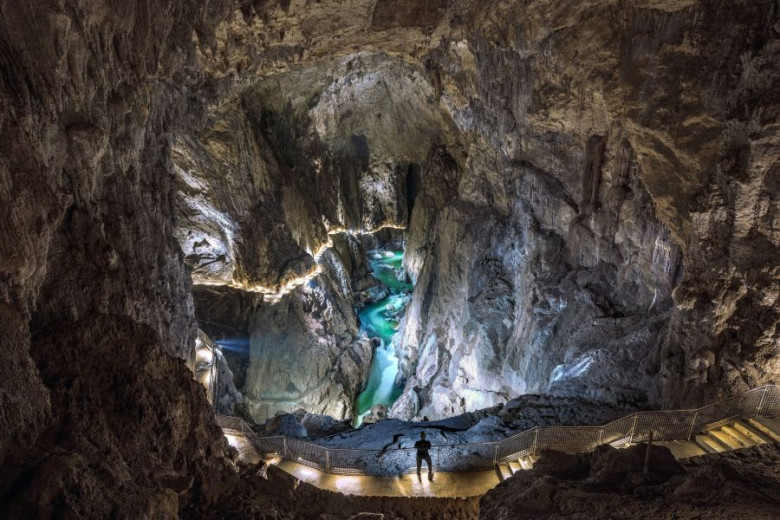A vast cave with a river and lit walkways reveals towering rock walls deep below.