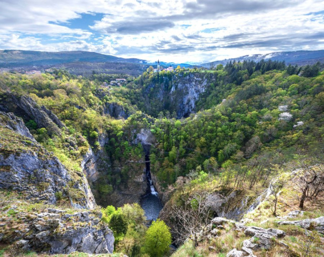 A green gorge with steep cliffs hides a waterfall, with a scattered settlement above.