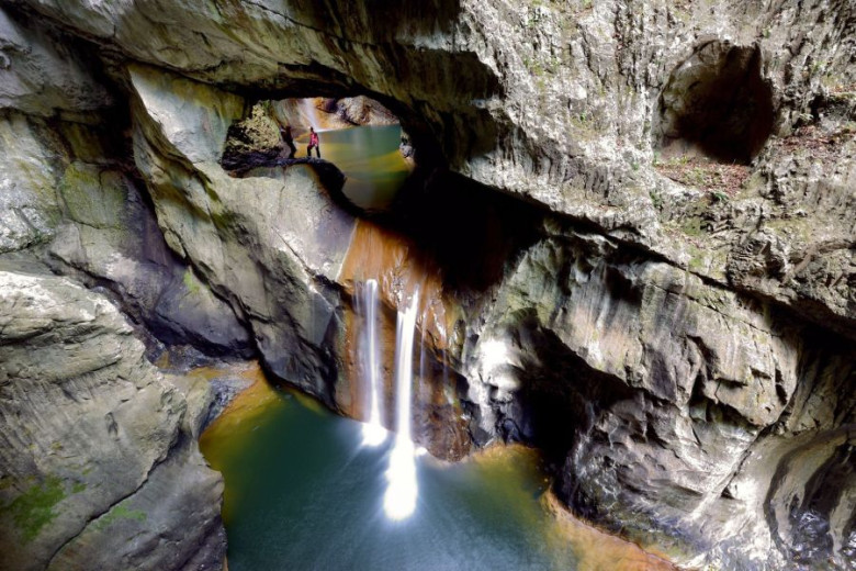 A tall waterfall drops into a green pool, with an explorer standing above on a natural arch.