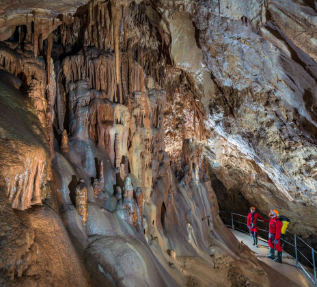 Cavers on a secured walkway observe speleothem formations rising throughout the spacious underground chamber.