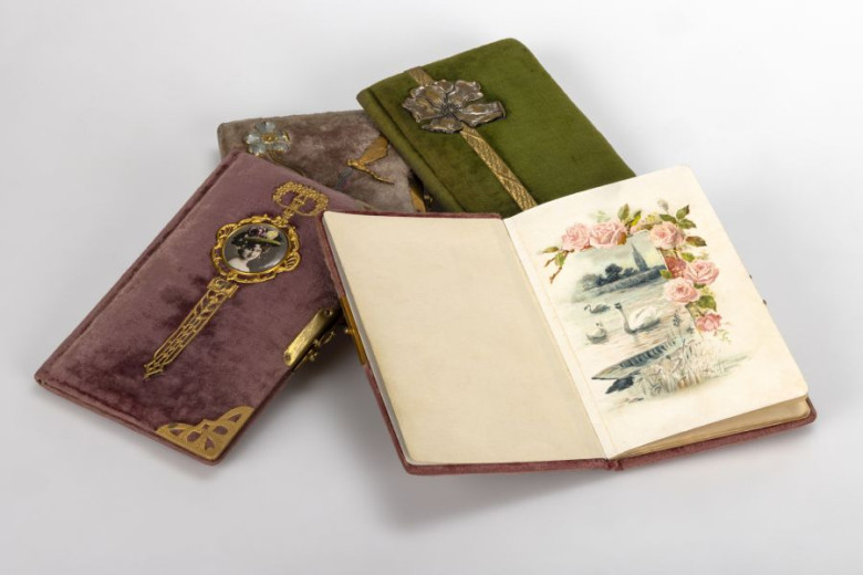Na mizi leži več okrašenih starih beležnic. Odprta prikazuje ilustracijo rožnatih vrtnic, vode in čolna. Oviti so v žametne vijolične in zelene platnice z bogatimi kovinskimi okraski.