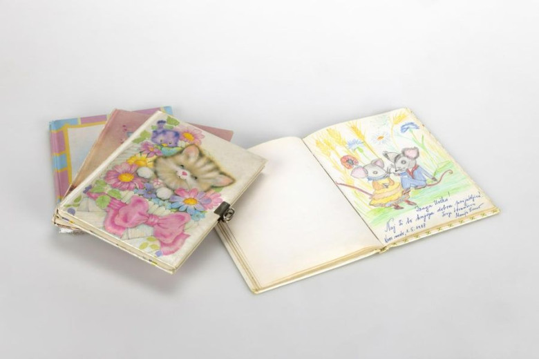 Several illustrated notebooks lie open and closed. One cover features a kitten surrounded by flowers, while an open book shows a colourful drawing of two mice holding umbrellas, accompanied by handwritten text.