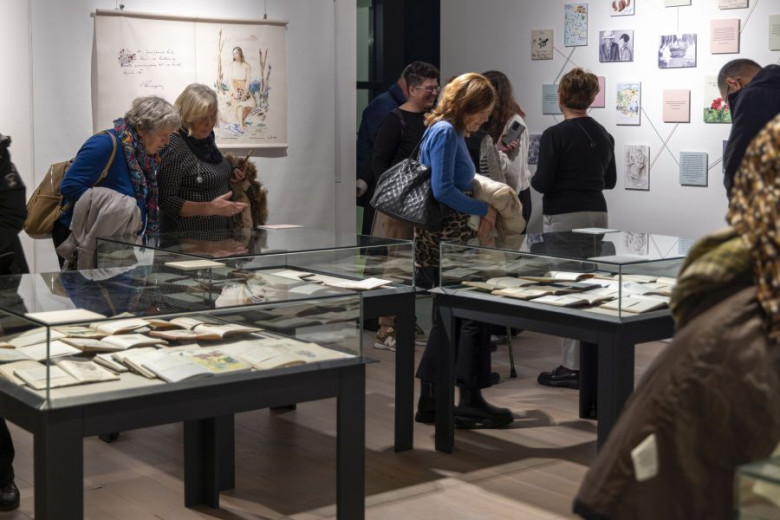 Obiskovalci si v galeriji ogledajo ilustrirane beležnice v steklenih vitrinah. Nekateri listajo po razstavljenih straneh, drugi opazujejo dela na steni. Levo visi večja ilustracija.