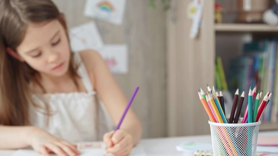 Otrok sedi za mizo in riše s škrlatnim svinčnikom. V ospredju je lonček z barvnimi svinčniki v ostrem fokusu, medtem ko sta otrok in ozadje z risbami mehko zamegljena.