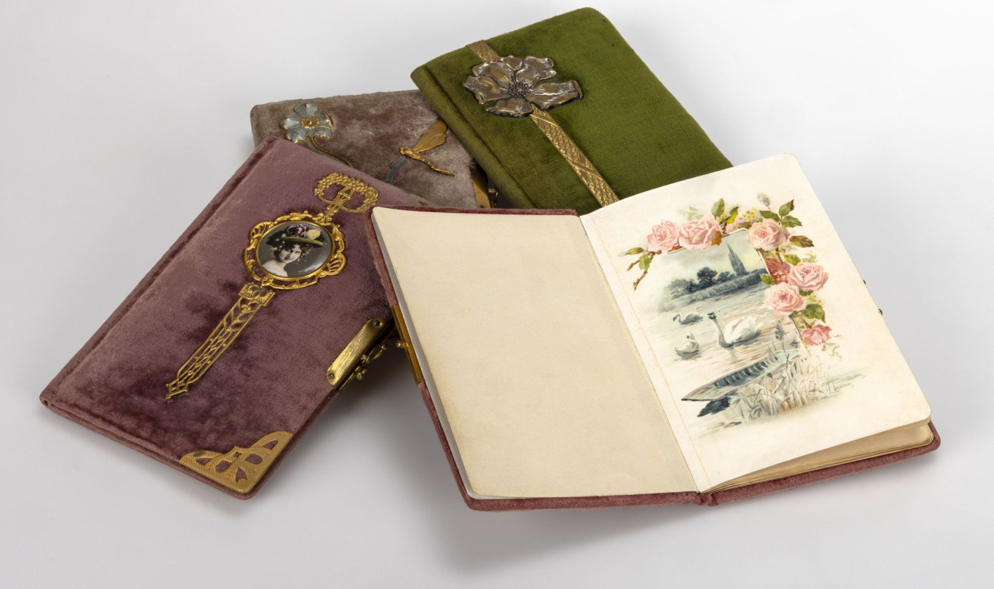 Several velvet-covered notebooks with ornate metal clasps lie arranged together. One is open, displaying a delicate illustration of pink roses above a lakeside scene with boats and birds.
