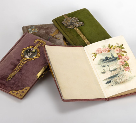 Several velvet-covered notebooks with ornate metal clasps lie arranged together. One is open, displaying a delicate illustration of pink roses above a lakeside scene with boats and birds.