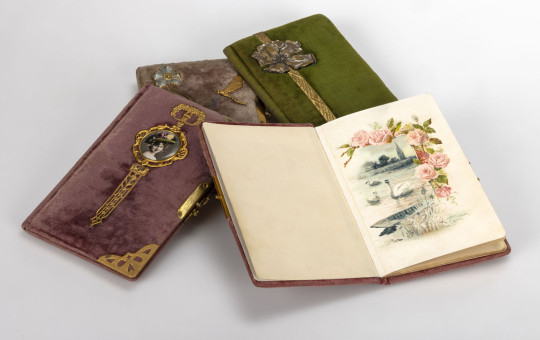 Several velvet-covered notebooks with ornate metal clasps lie arranged together. One is open, displaying a delicate illustration of pink roses above a lakeside scene with boats and birds.
