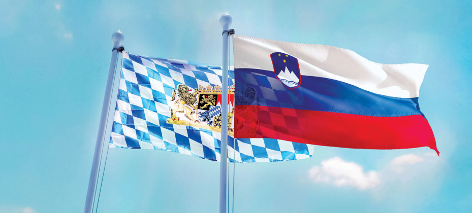 The Slovenian and Bavarian flags wave on flagpoles against a blue sky with clouds.