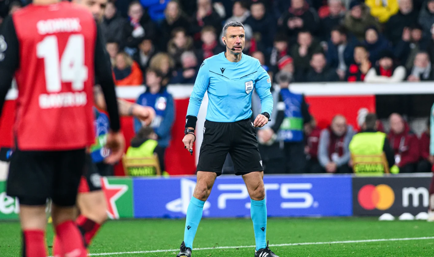 A football referee stands on the pitch during a match. The stands in the background are filled with spectators.