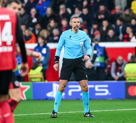 A football referee stands on the pitch during a match. The stands in the background are filled with spectators.