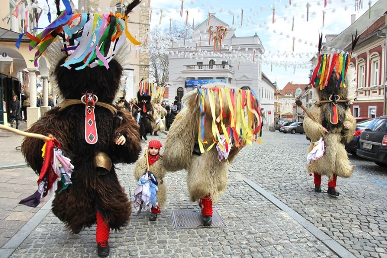 Kurents in traditional carnival costumes with bells and colorful ribbons dancing through a town street during a carnival parade.