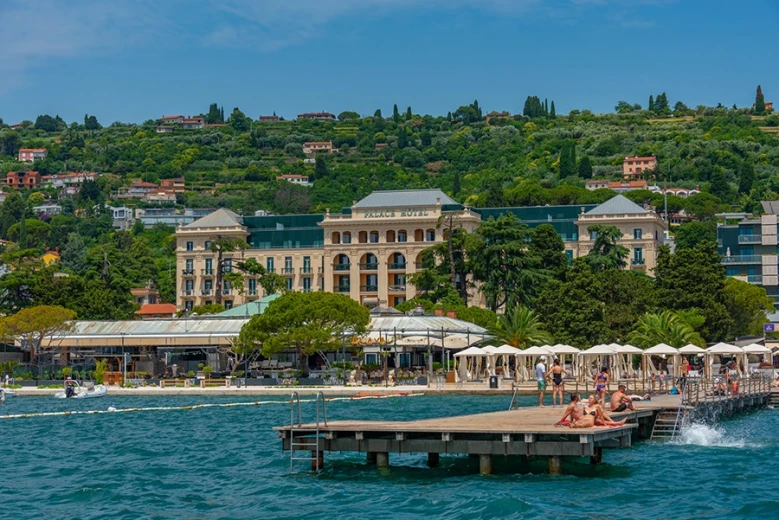 Swimmers relaxing on a pier by the sea in front of the Palace Hotel in Portorož, with a hillside of villas and trees rising in the background.