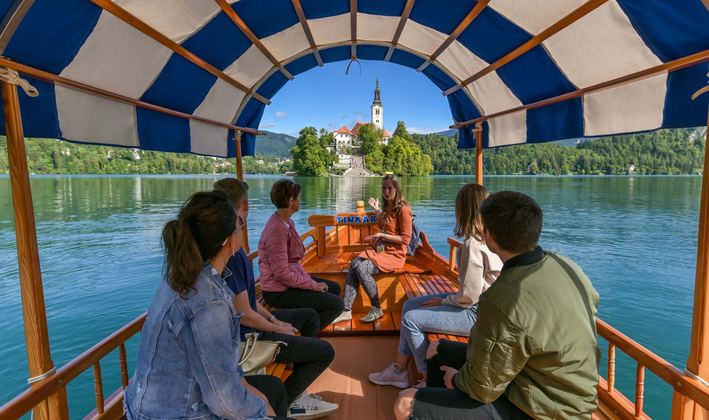 Skupina ljudi se vozi s tradicionalnim blejskim čolnom, pletno, proti otoku s cerkvijo na Blejskem jezeru ob sončnem dnevu.