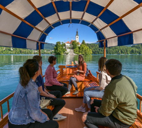 A group of people riding a traditional Bled pletna boat towards the island with the church on Lake Bled on a sunny day.