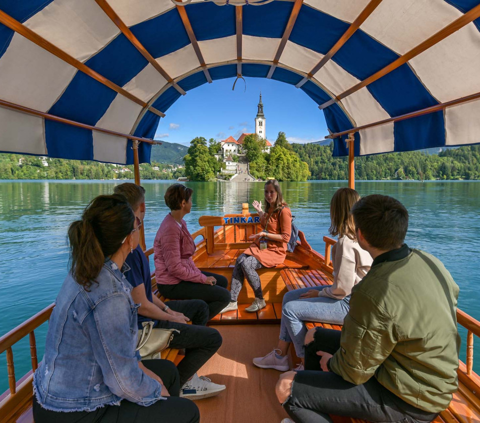 A group of people riding a traditional Bled pletna boat towards the island with the church on Lake Bled on a sunny day.