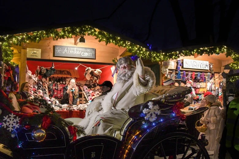 Grandfather Frost greets visitors from a festively decorated sleigh at a Christmas market, surrounded by wooden stalls and lights.