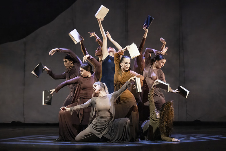 A group of dancers performing a contemporary dance piece, holding open books in their hands.