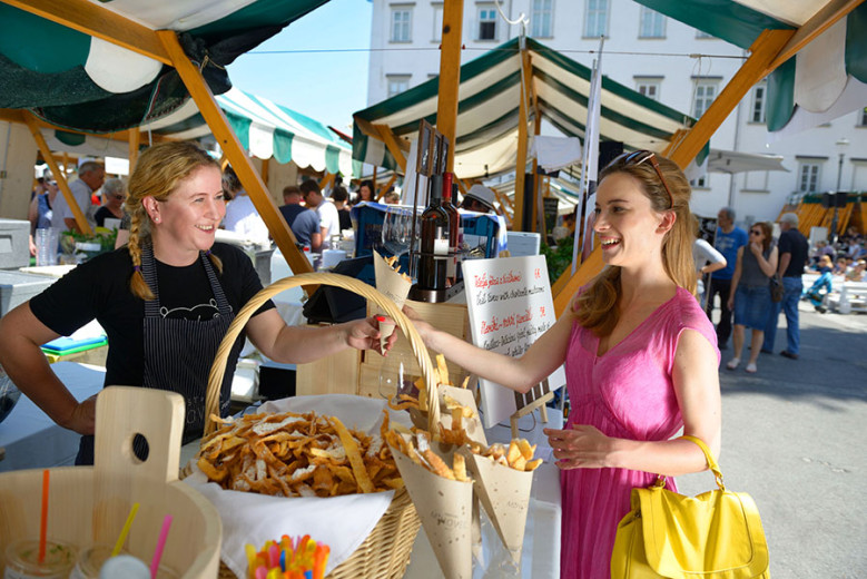 A vendor at an open-air market offers food in a paper cone to a visitor, with stalls and people in the background on a sunny day.