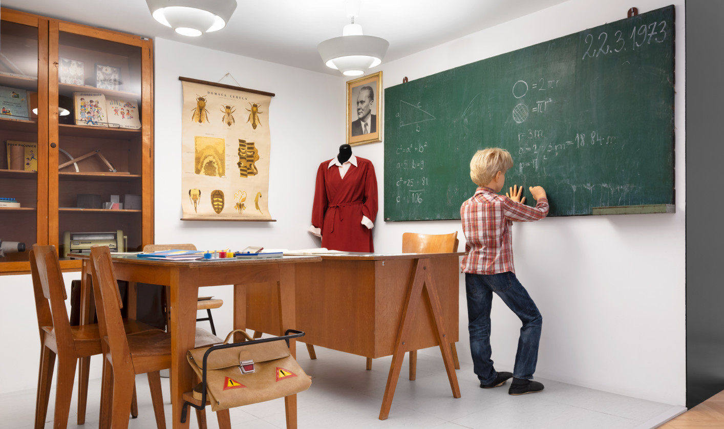 A boy in a reconstructed 1970s classroom writes mathematical formulas on the blackboard, surrounded by wooden furniture, teaching materials, and vintage school items.