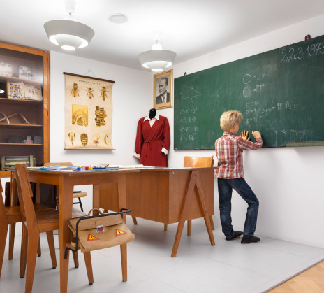 A boy in a reconstructed 1970s classroom writes mathematical formulas on the blackboard, surrounded by wooden furniture, teaching materials, and vintage school items.