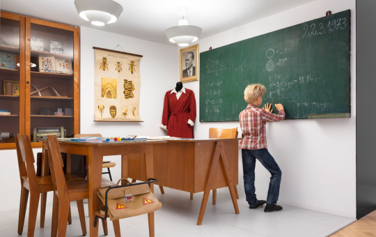 A boy in a reconstructed 1970s classroom writes mathematical formulas on the blackboard, surrounded by wooden furniture, teaching materials, and vintage school items.